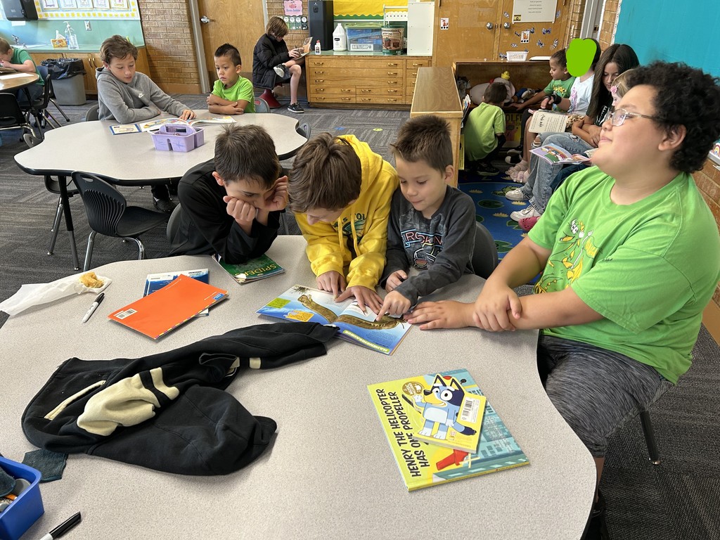 An indoor photo of four older students sitting around a gray table, looking at an open picture book together. One student in a bright green shirt is smiling on the right, while the other three students are clustered closely around the book in the center. Other students are visible reading in the background.