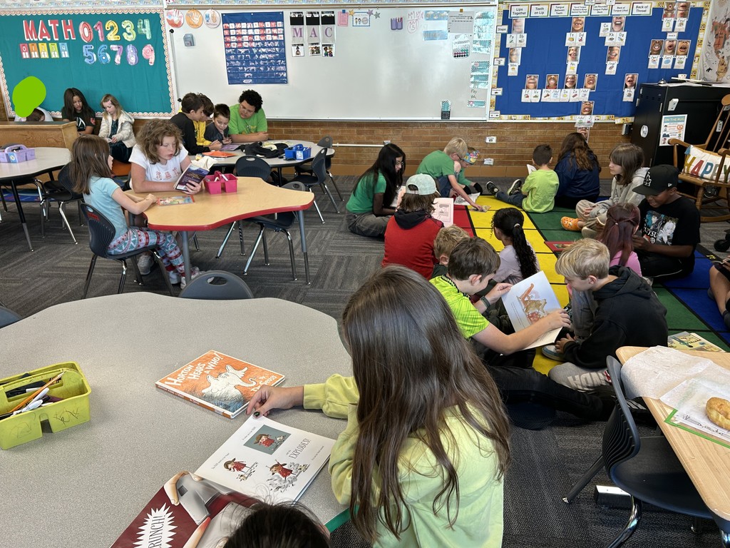 A wide, indoor photo of a busy elementary classroom filled with students. Students are paired up and scattered around the room, sitting at tables or on the floor, reading books together during a reading buddies session.