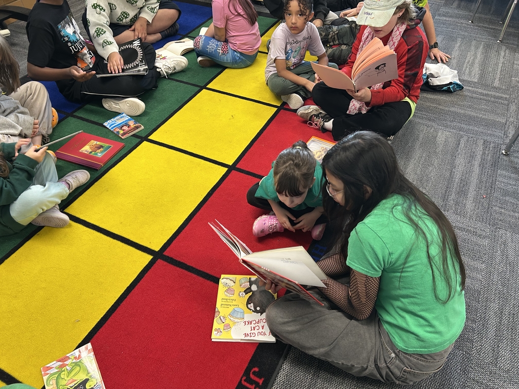 A close-up, overhead photo of a group of students sitting on a colorful carpet. An older student in a green shirt is reading a book aloud to a young student, their kindergarten reading buddy.