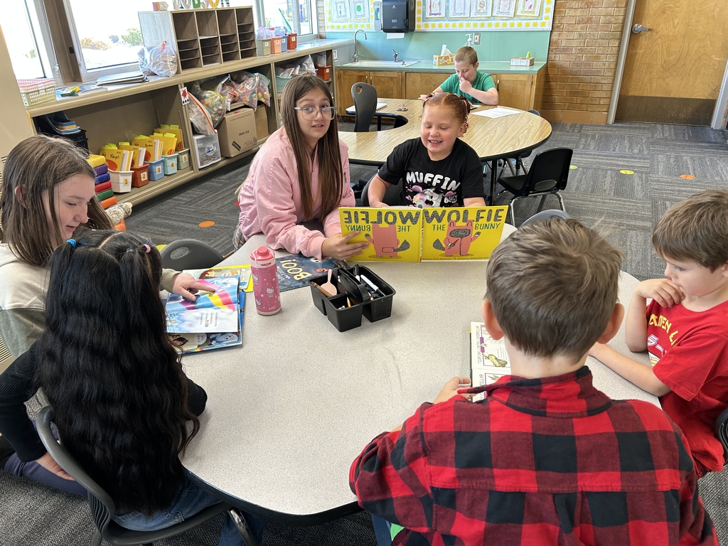 An overhead photo of two older students sitting across from three younger students around a hexagonal table in a classroom. The students are engaged in a reading buddy session, looking at and discussing open books on the table.