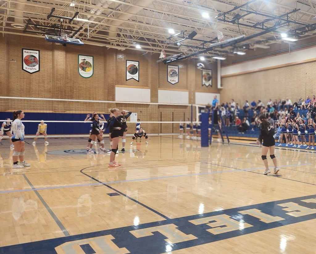 Fairfield's player is serving the ball.  One of the girls is showing numbers with her hand to indicate where the ball should be served.  Fairfield and Centerville players are watching, ready to play.  The crowd is seated in the bleachers to the right side of the picture.