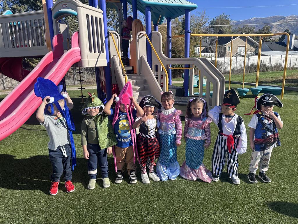 a group of 8 young students dressed in pirate, mermaid, squid and dinosaur costume gathered in front of a play structure,.