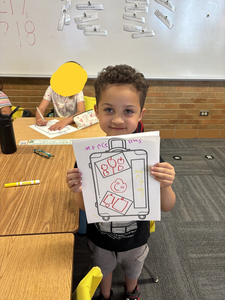 A curly-haired boy in a classroom, wearing a black shirt and gray shorts, looks directly at the camera while proudly holding up a sheet of paper. The paper features his colored-in drawing of a Culture Suitcase decorated with stickers and symbols representing his heritage. Another student is partially visible at a desk in the background.