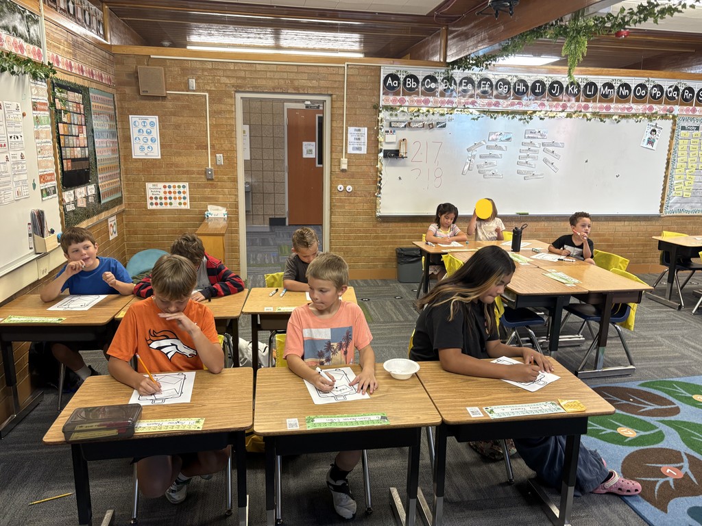 Eight elementary-aged students are shown seated at individual wooden desks in a classroom, working on a drawing activity. The students are concentrated on coloring paper templates of suitcases, which is the Culture Suitcase project. The classroom features a whiteboard with math problems, an alphabet banner, and brick walls decorated with educational posters.