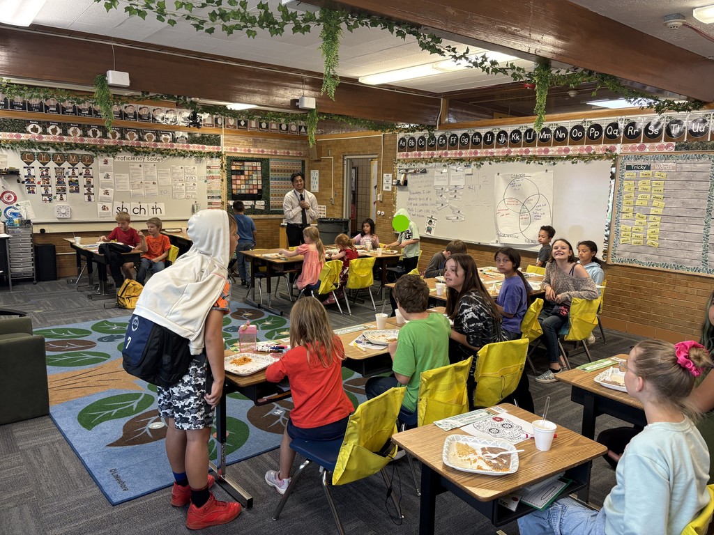 A wide shot of a busy classroom during the Multicultural Club's Hispanic Heritage Month celebration. Students are seated at tables with yellow chairs, many looking towards the teacher or club leader standing near a whiteboard. Plates of food, including rice and beans, are visible on the desks, and there's a colorful rug in the foreground.