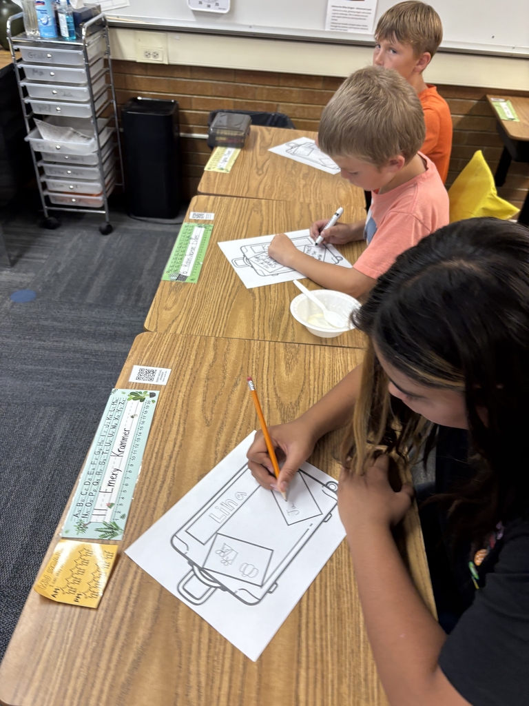 Three students are sitting at desks drawing. The student in the foreground, with long dark hair, is intently coloring her paper with a pencil. Two other students, both blonde boys, are seated further down the table, also focused on their drawings. A small white bowl, holding ice cream, sits on the table next to one of the students.
