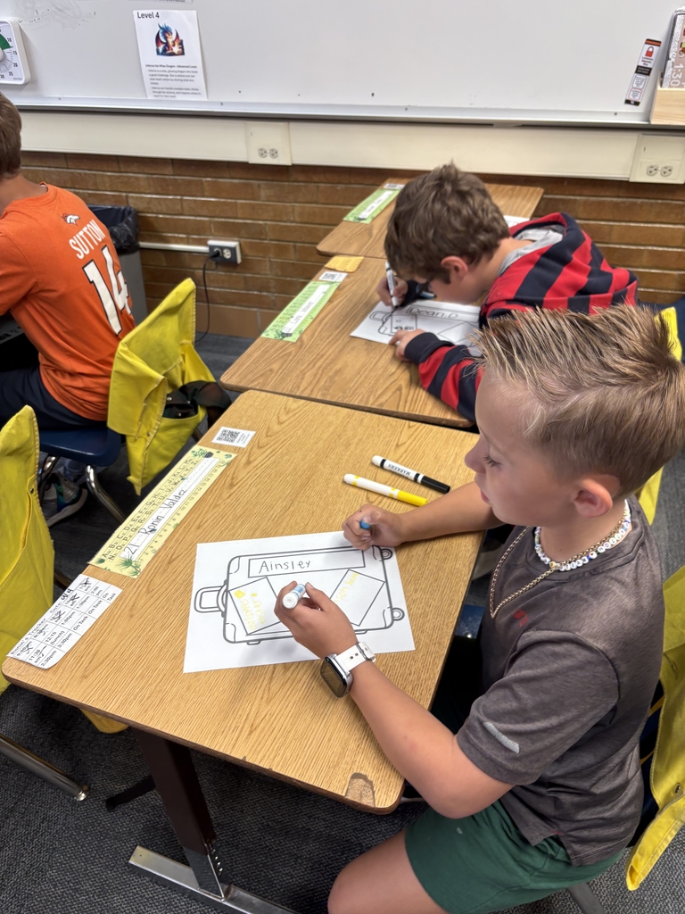 A close-up shot of a young student with blonde hair, wearing a dark gray shirt and a beaded necklace, working on a drawing labeled "Ainsley." The student is using a marker to color in the design of a Culture Suitcase on a desk. Two other students are visible working at desks behind him.