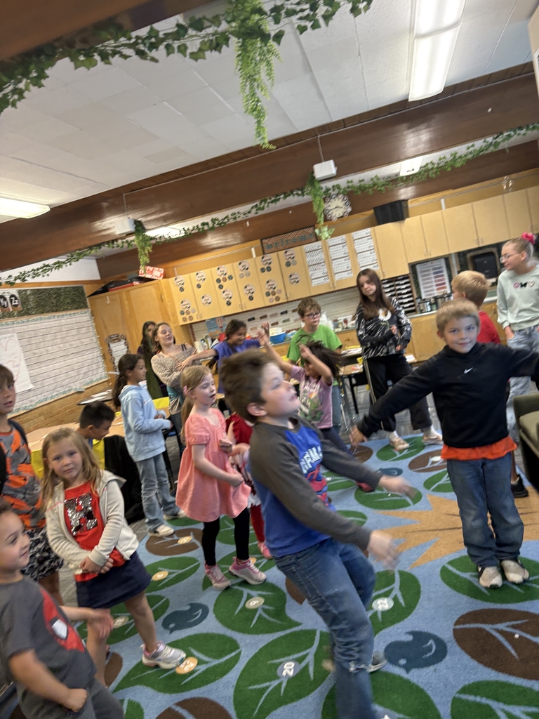A group of children are active and smiling in a classroom setting, dancing or playing a movement game on a blue rug with a green leaf pattern. Their arms and bodies are in motion, suggesting an energetic game like freeze dance or a salsa lesson, which was part of the Hispanic Heritage Month celebration.