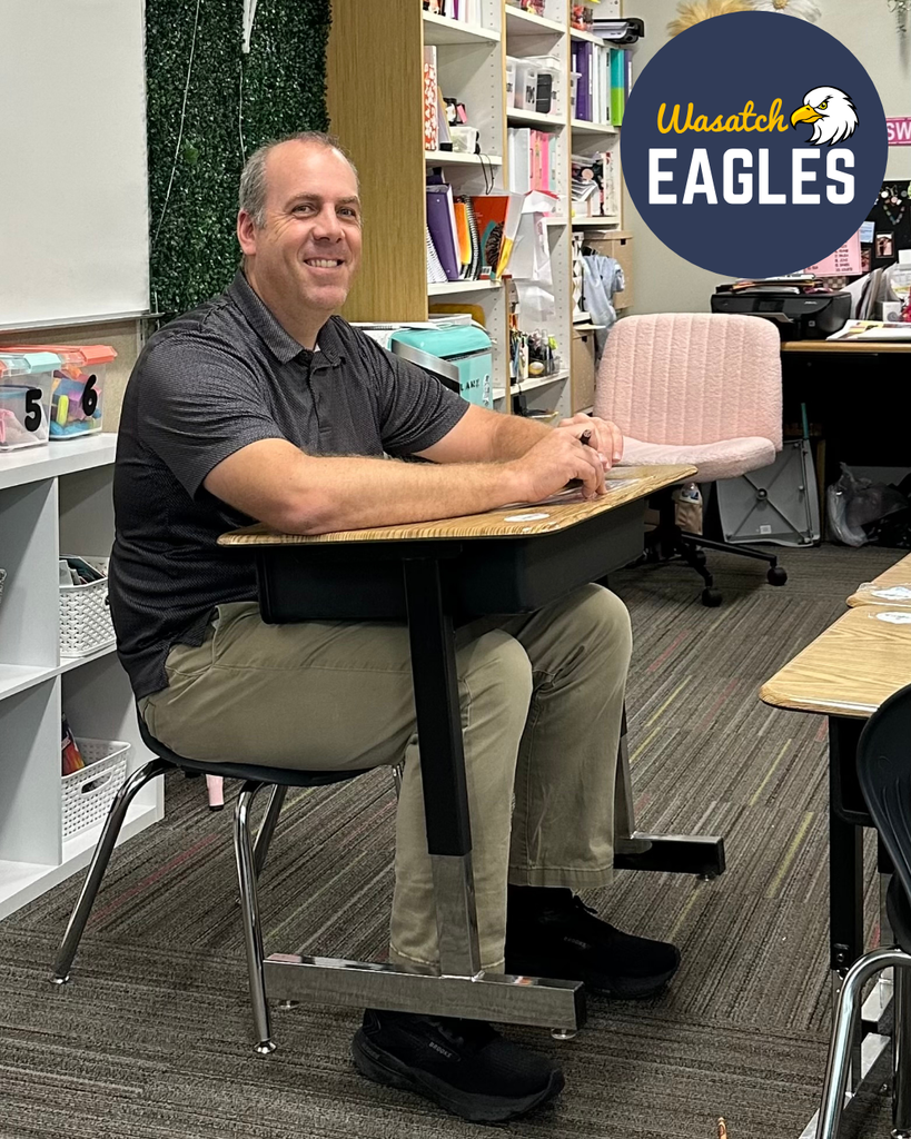 A tall teacher sitting at a small student's desk.