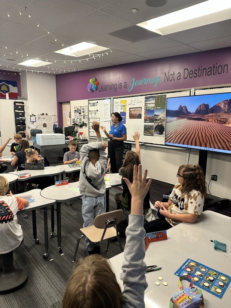students raising hands while a woman presents at the front of the room