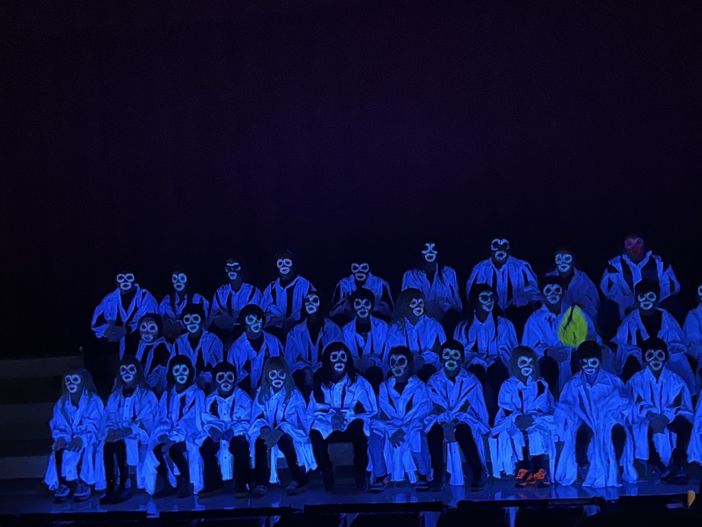 A large group of students in a dark room, dressed in glow in the dark clothing with their faces painted, singing halloween songs
