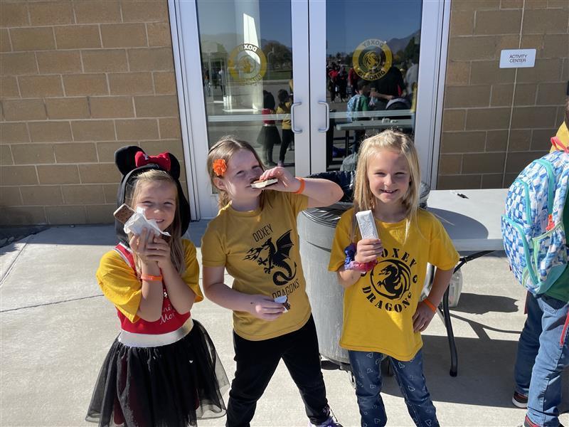 Three young girls smiling and eating treats, an ice cream sandwich, outside of a school building. Two are wearing yellow "Doxey Dragons" t-shirt