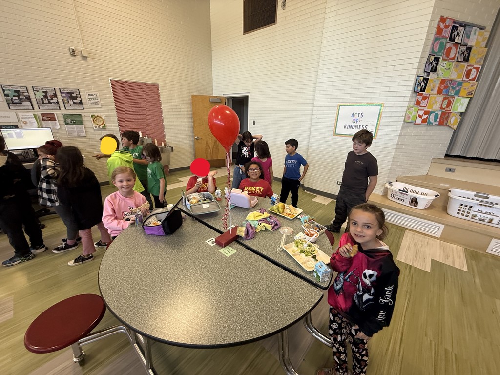A group of elementary school students gathered around a circular table in a school cafeteria for a special lunch. Five children are visible at the table with lunch trays and a large red balloon. Other students are standing nearby. A sign on the wall behind them reads "Acts of Kindness."