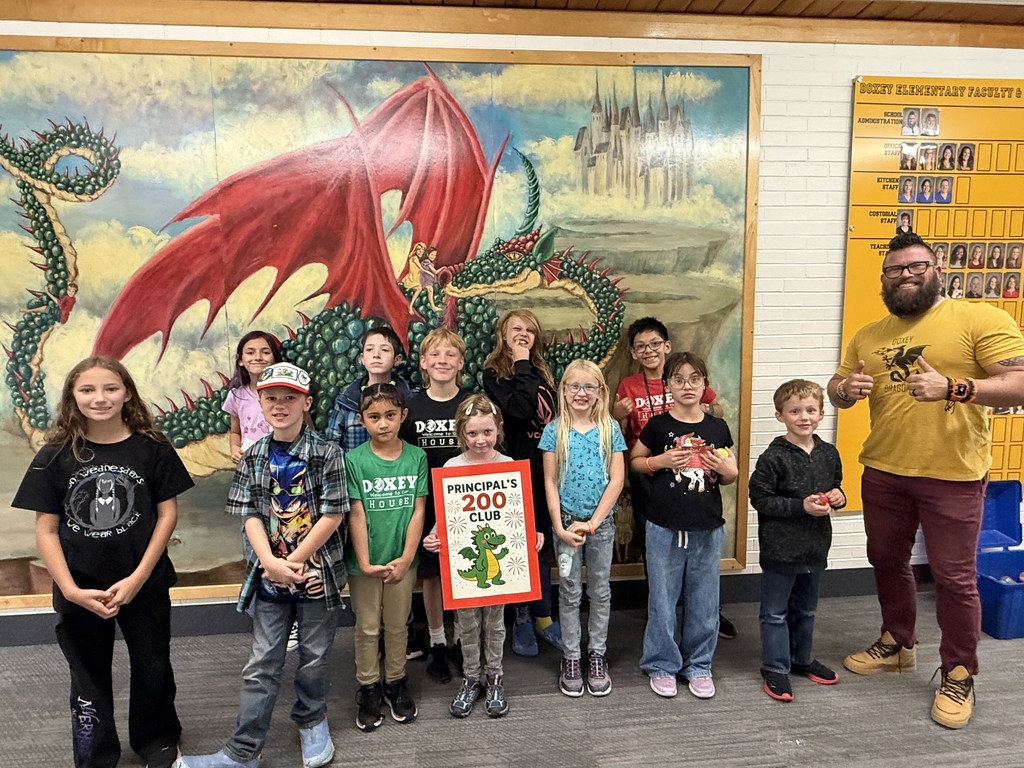 A group of approximately ten elementary school students and one adult man posing for a photo in front of a large mural depicting a red-winged dragon and a castle. The students are holding a sign that reads "Principal's 200 Club" with a drawing of a green dragon. The principal Hansen on the right, wearing a yellow Doxey Dragons shirt, gives a thumbs up.