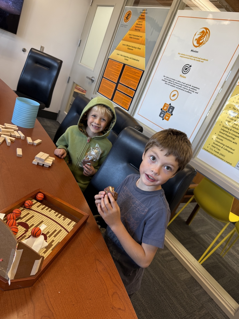 Two young elementary school boys smiling while sitting at a conference table in an office, enjoying chocolate treats. Wooden blocks (like Jenga) and a miniature basketball game are scattered on the table. A poster detailing the school's mission and vision is visible in the background.