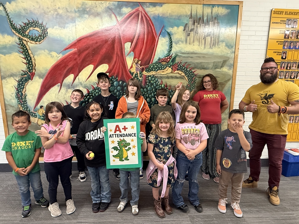 A diverse group of about twelve elementary school students and two adults posing for a photo in front of a large mural of a green and red dragon and a castle. The students are holding a large green sign with a red border that says "A+ Attendance" and features a drawing of a small green dragon. The adult man, Principal Hansen on the right, wearing a yellow Doxey Dragons shirt, is giving two thumbs up.