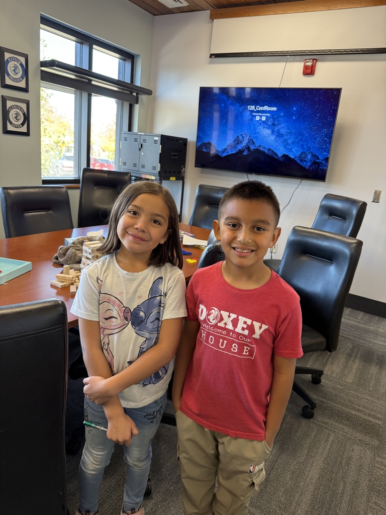 Two elementary-age children, a girl and a boy, stand side-by-side and smile for the camera in an office setting. They are in a conference room with a large dark wood table, leather chairs, and a large wall-mounted screen displaying a starry night sky image. The boy is wearing a red shirt that says "Doxey Welcome to Our House."