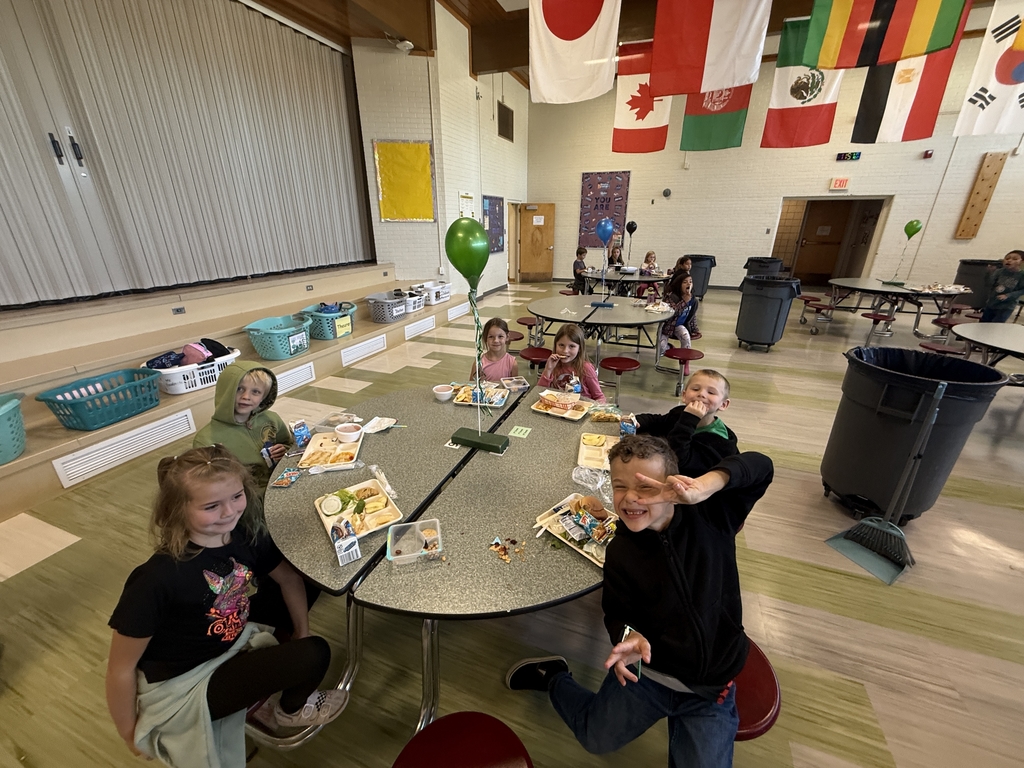 A group of six young, smiling students sitting around a round table in a school cafeteria, enjoying a "house lunch" party. Lunch trays are visible, and a green balloon is tied to the table. International flags hang from the ceiling above the dining area.