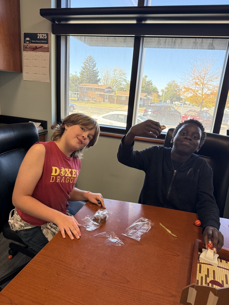 Two elementary school students sitting at a large wooden desk in front of a sunny window, enjoying treats during a party. The girl on the left wears a maroon "Doxey Dragon" tank top and is smiling. The boy on the right is holding up a small snack and grinning.