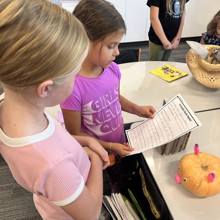 a young girl reads a paper to another girl with pumpkin in front