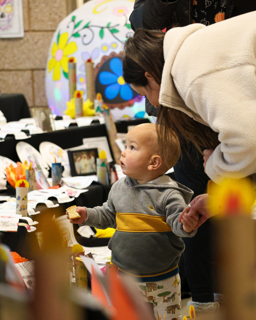 A child looks at the ofrenda with a parent while holding cake.