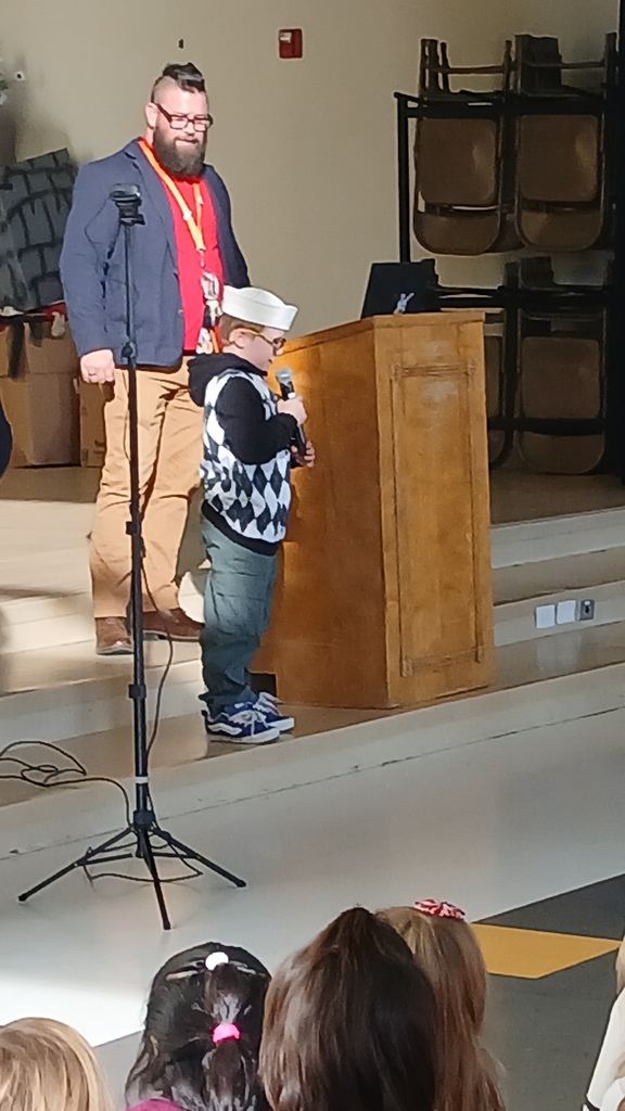 Student wearing navy hat standing in front of the assembly with principal Hansen sharing his experience with his grandpa that served and passed away.