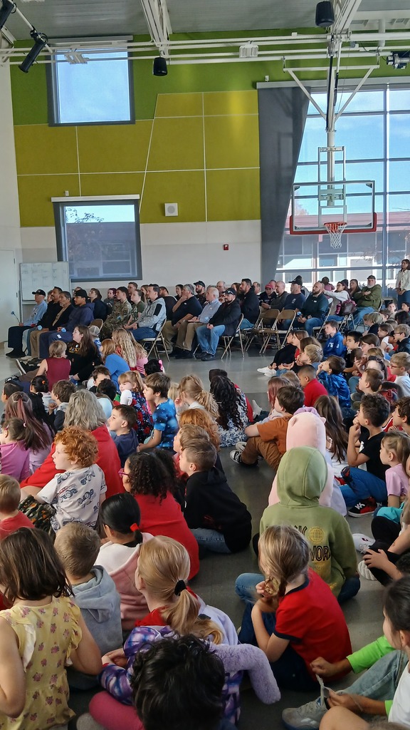 A wide shot of a very fully gym. students are sitting on the floor while the veterans being honored are seated in folding chairs.