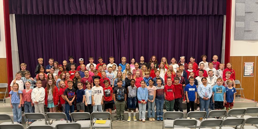 Fifth grade students standing in rows on stage steps with a purple curtain backdrop.