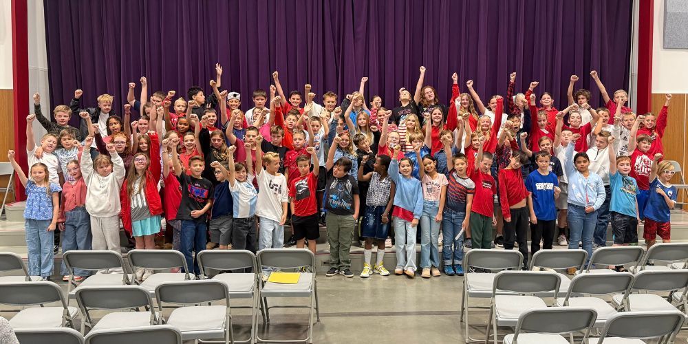 Fifth grade students standing in rows on stage steps with a purple curtain backdrop. They are all holding a fist in the air cheering.