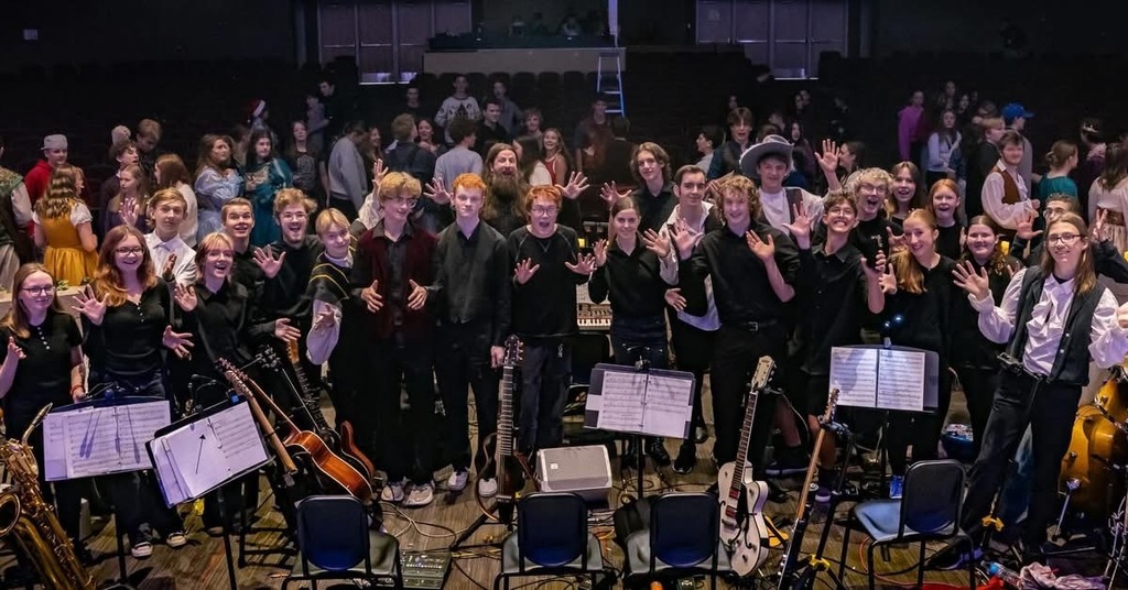 A large group of smiling students, many dressed in black performance clothes, stand on stage in front of music stands and instruments, waving at the camera with an audience of other students gathered in the background.