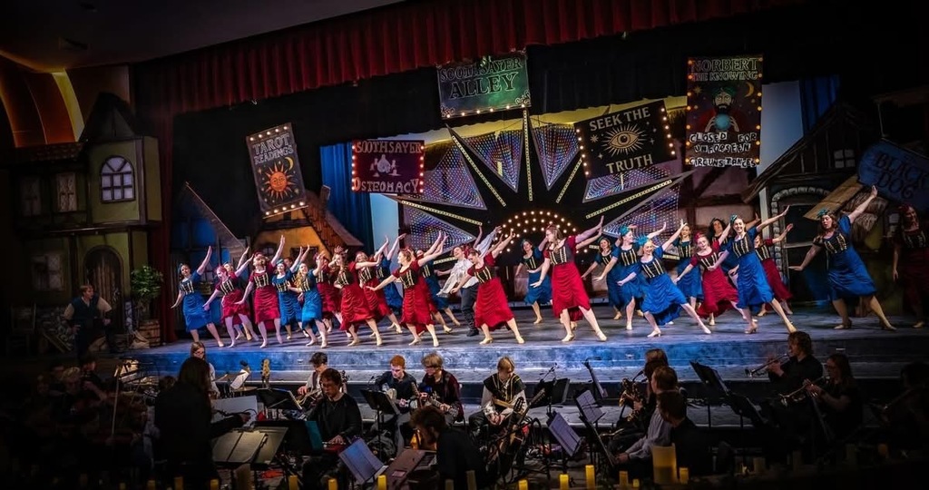 A large musical theatre production with a line of dancers in matching red and blue costumes performing across a brightly lit stage, backed by colorful carnival-style signs, while a live orchestra plays in the pit below.