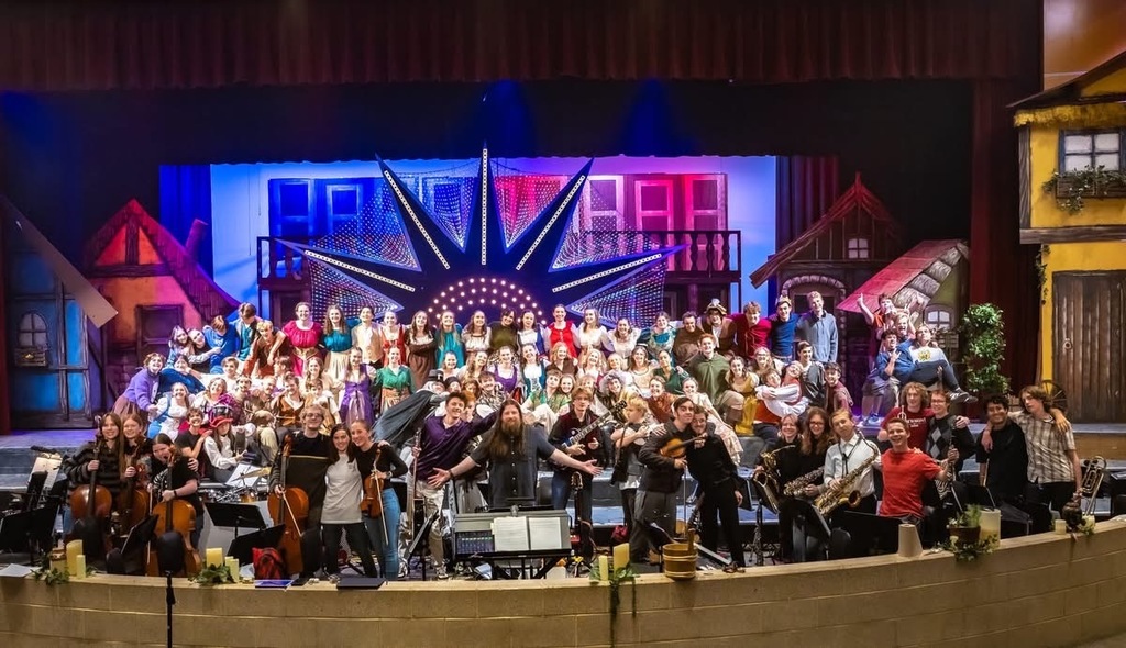 Large group portrait of a school musical’s full cast and orchestra posed on a brightly lit stage with colorful village scenery and a large starburst light backdrop.