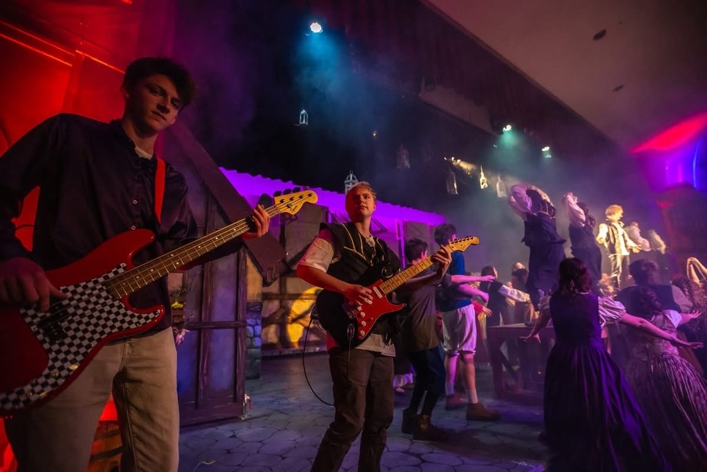 A stage performance with dramatic colored lighting, showing two young musicians in the foreground playing electric guitars while a group of costumed actors dances and performs in the smoky, brightly lit background.