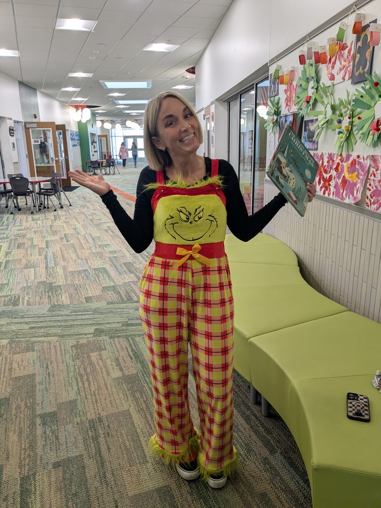 Teacher wearing a Grinch outfit smiling at the camera holding a book. 