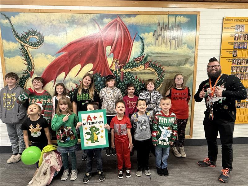A large group of Doxey Elementary students, the A+ Attendance winners, standing with a teacher in front of the school's dragon mural. The students are holding a sign that says "A+ Attendance" and many are wearing holiday-themed clothing.