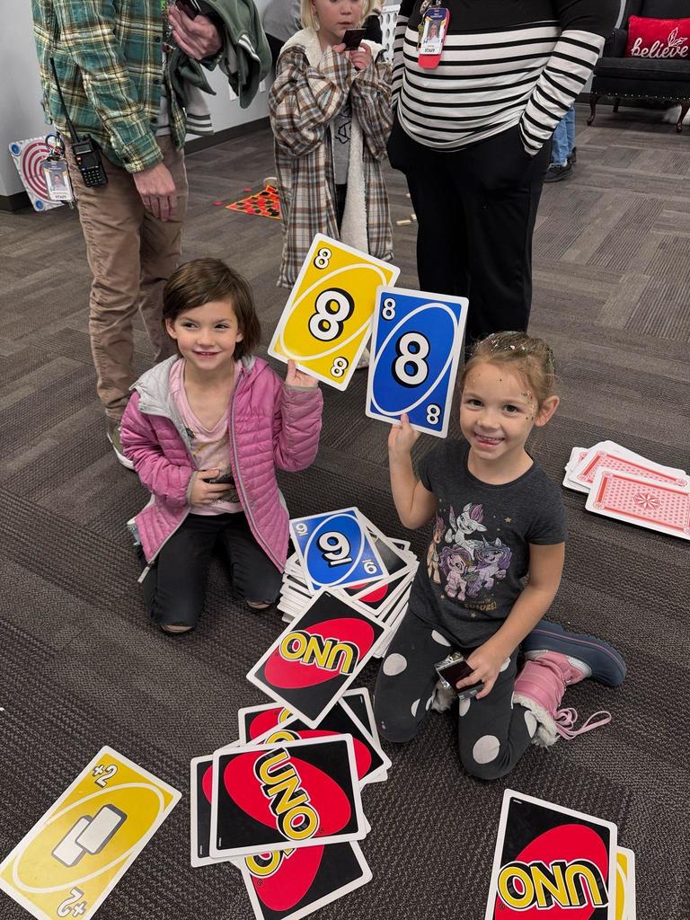 Two young Doxey Elementary girls kneeling on the floor, holding up large UNO cards and surrounded by a pile of colorful UNO cards