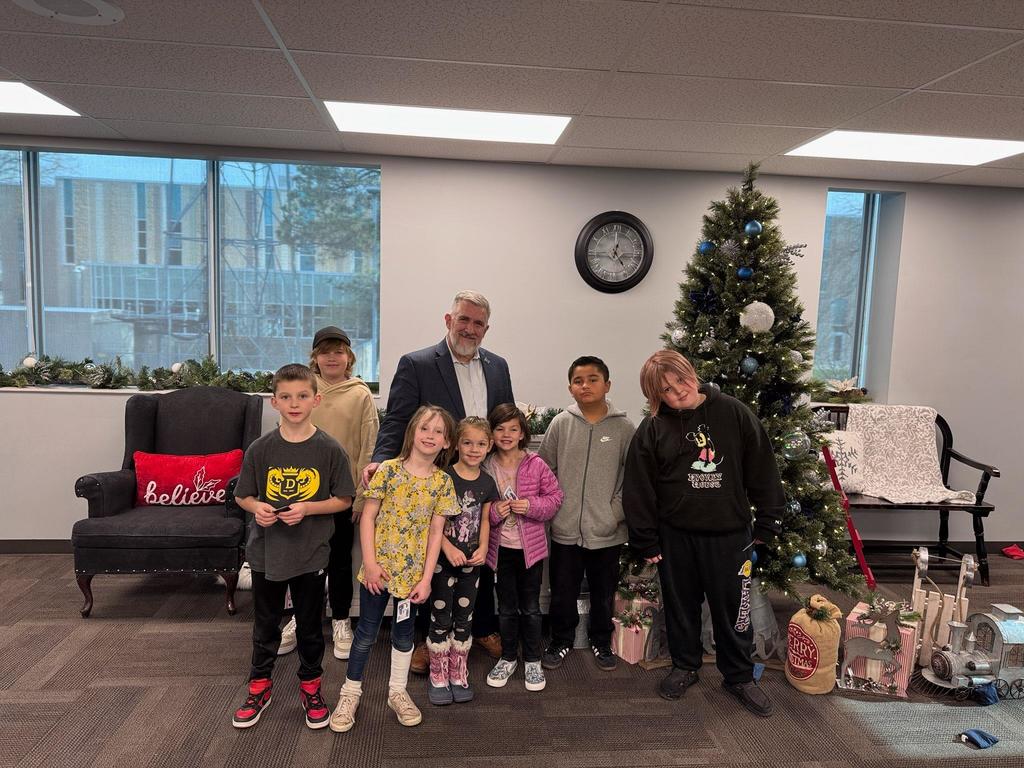 Six young Doxey Elementary students and Clearfield City Mayor Mark Shepherd stand together in an office setting with a decorated Christmas tree.