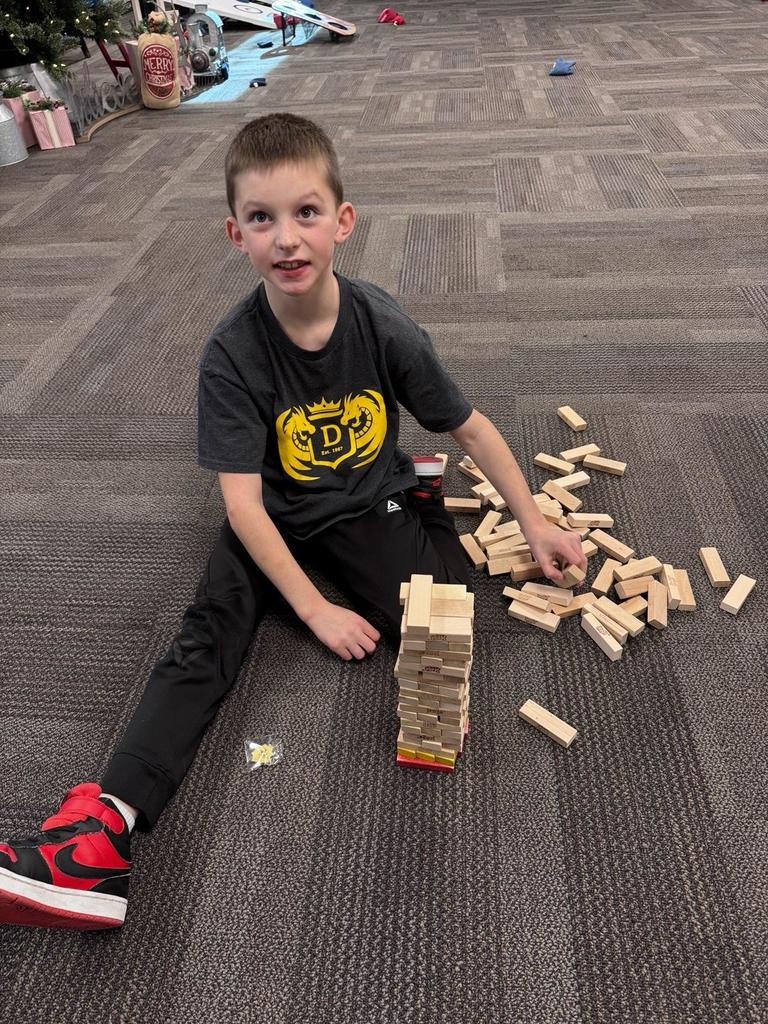 A Doxey Elementary student sitting on the floor, playing a game of Jenga with a partially built tower and scattered wooden blocks. He is wearing a grey Doxey Dragons shirt.