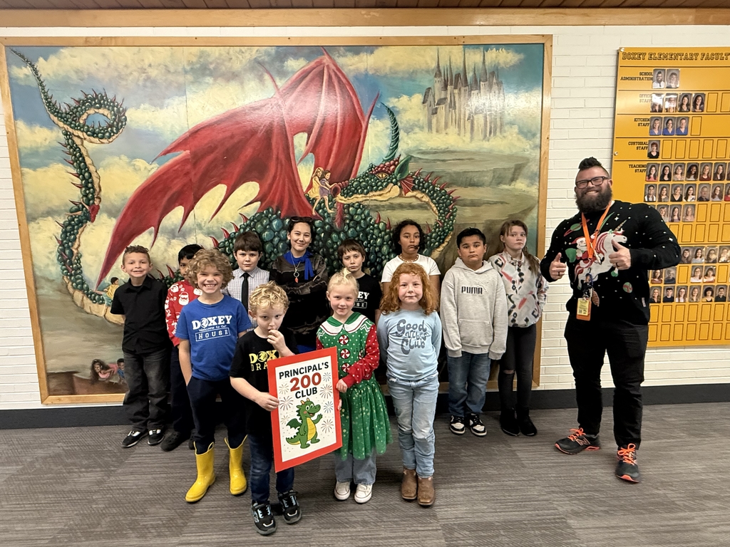 Doxey Elementary Principal's 200 Club winners of the week (ten children) posing with their principal in front of a mural depicting a red and green dragon. The principal, wearing a festive sweater, is giving a thumbs-up.