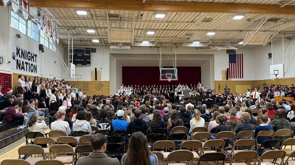 image of students in band, orchestra and choir performing at an assembly. spectators are in the audience in chairs and on the bleachers.