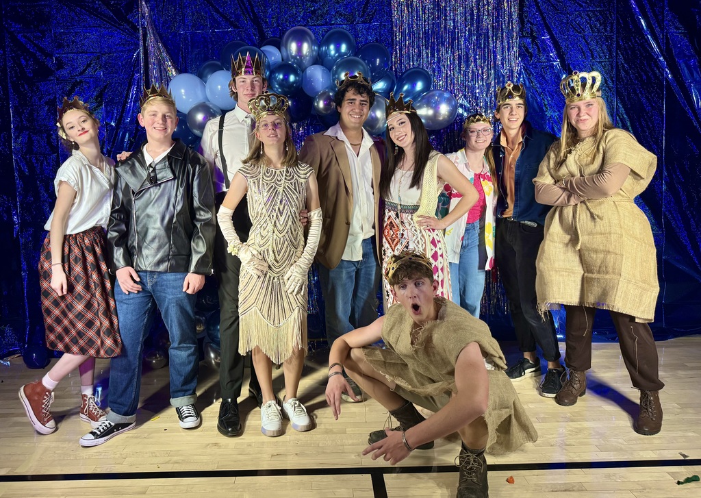 A group of ten teens/young adults pose on a gym floor in front of a shiny blue photo backdrop and a cluster of blue and silver balloons; most wear gold crowns and themed costumes (including a leather jacket, suspenders, a gold flapper-style dress with long gloves, patterned dresses, and burlap tunics). One person crouches in the front with a surprised open-mouthed expression while the others stand smiling and posing.