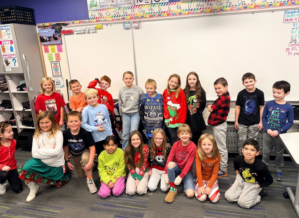 Elementary students smiling and posing together in a classroom, wearing festive holiday clothing in front of a whiteboard