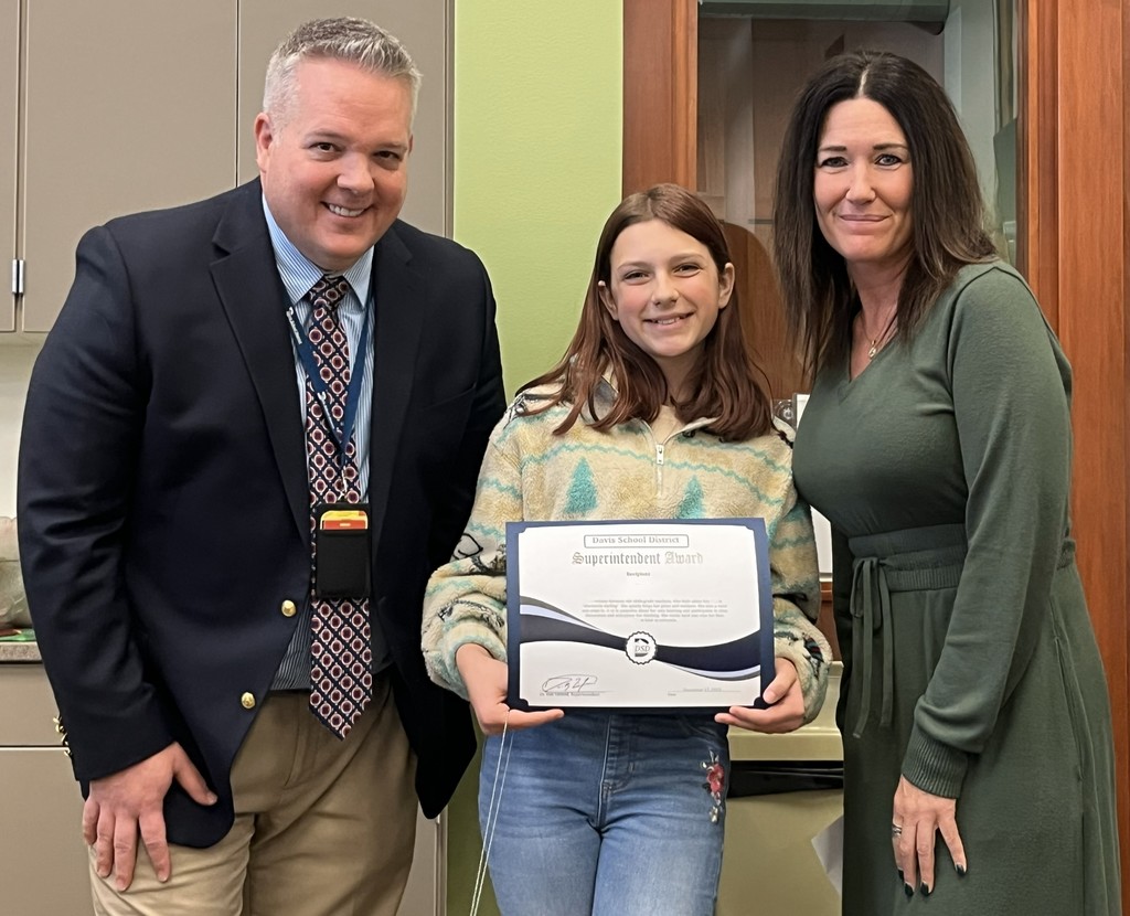 A student stands smiling while holding a Superintendent Award certificate, flanked by the superintendent and a female staff member in an office setting.