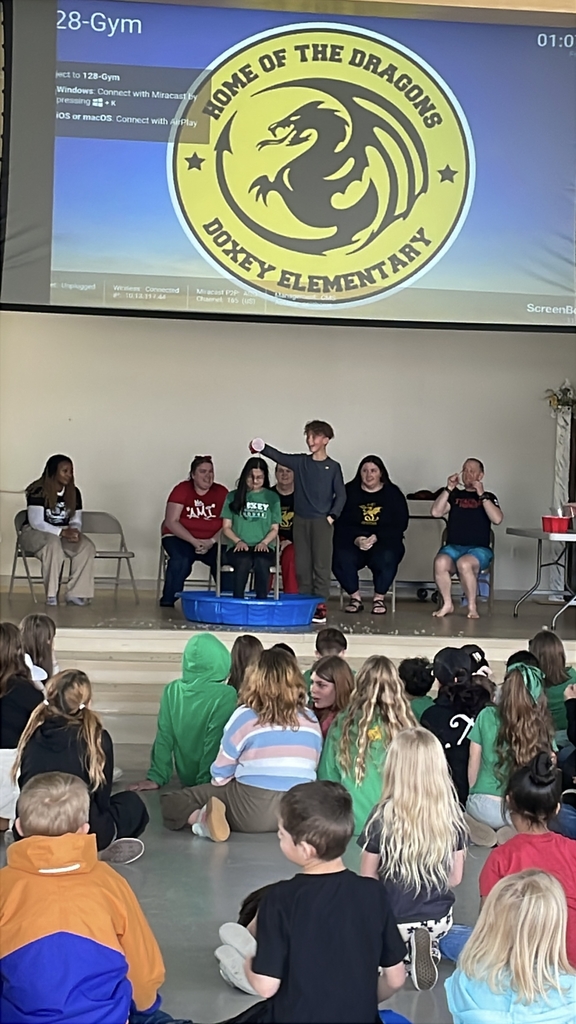 A teacher sitting in a blue kiddie pool on stage, completely drenched as a student pours a large bucket of water over them. The school's "Doxey Elementary" logo is visible on the screen behind them.