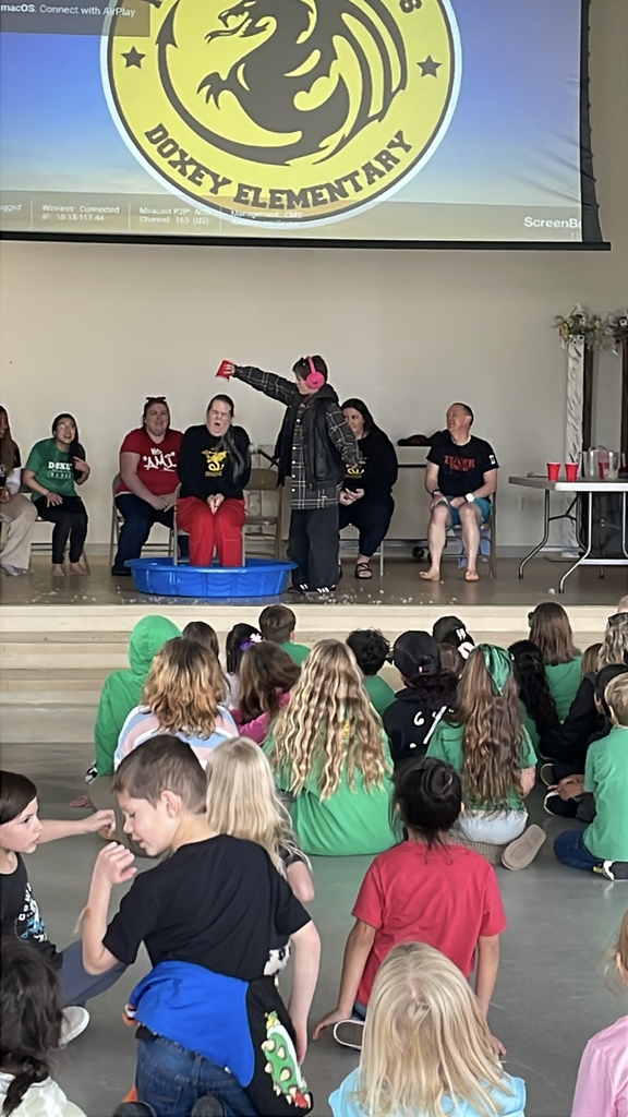 An action shot of a student pouring ice water onto a teacher who is sitting in a blue pool. You can see the ice cubes and water mid-air, with fellow teachers in the background cheering and smiling.