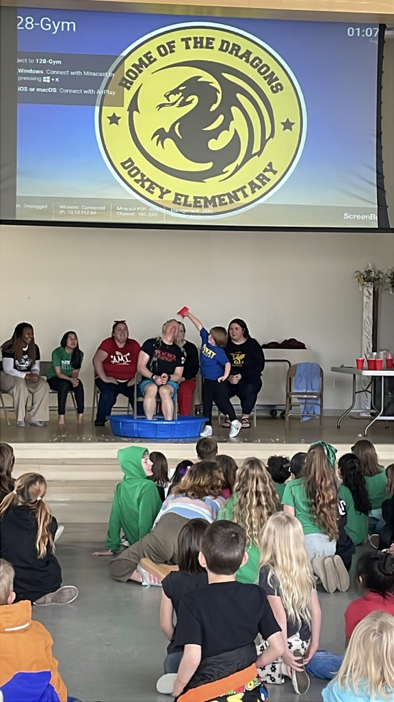 A close-up of a student in a blue shirt carefully tipping a silver bucket of ice and water over a teacher's head. The teacher is bracing for the cold, and water is splashing everywhere.