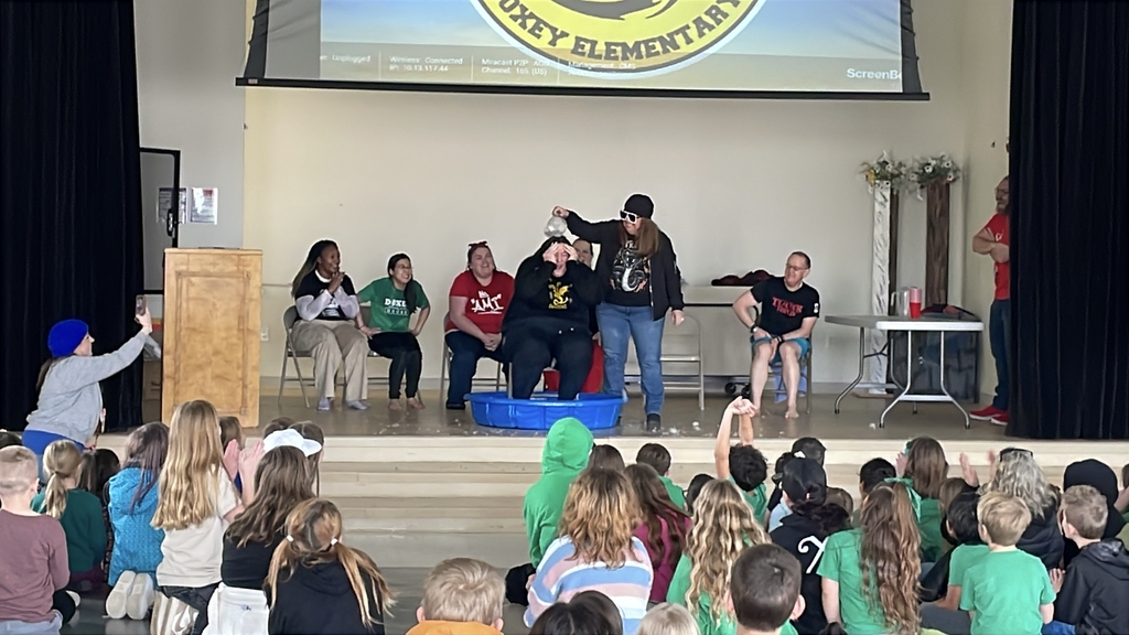 A wide shot of the Doxey Elementary stage during an assembly. A student in a black jacket and sunglasses pours a silver bucket of ice water over a teacher sitting in a blue plastic pool while other teachers watch and laugh.