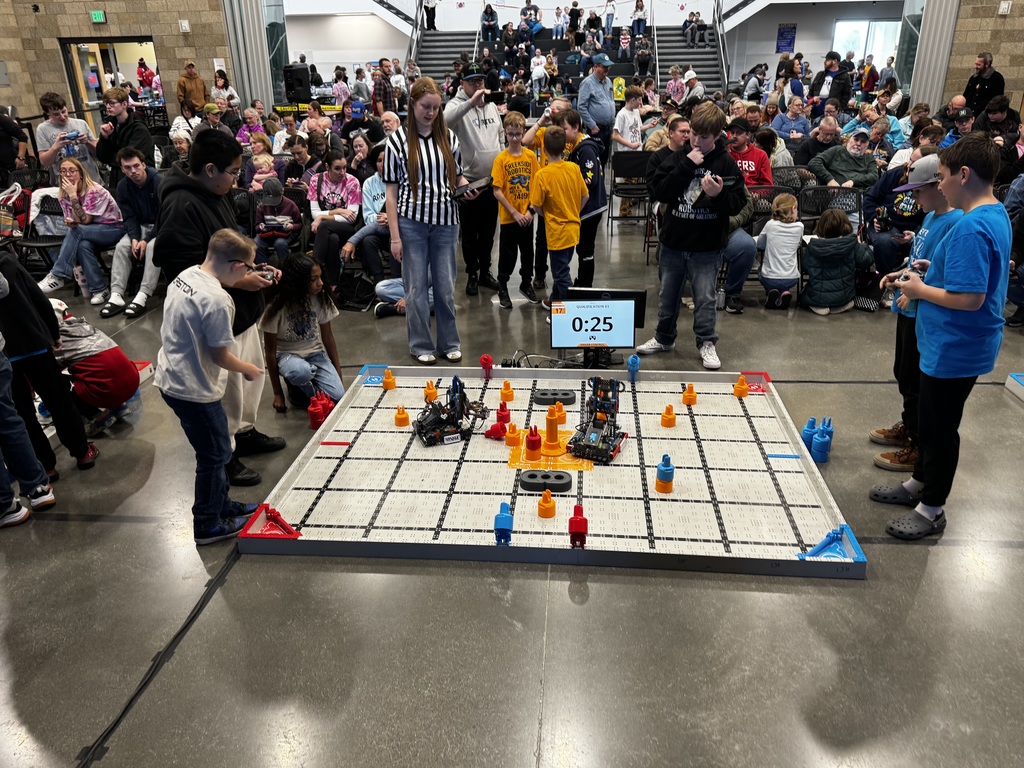 Students and Parents fill the gym to watch the VEX robotics competition.  Students are in the forefront  of the gym working together to complete their task.