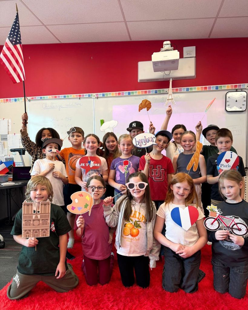 A group of elementary students stand and kneel together in a classroom holding French-themed photo props such as baguettes, berets, the Eiffel Tower, hearts, and speech bubbles. An American flag is visible in the classroom, and students are smiling and posing for the photo.
