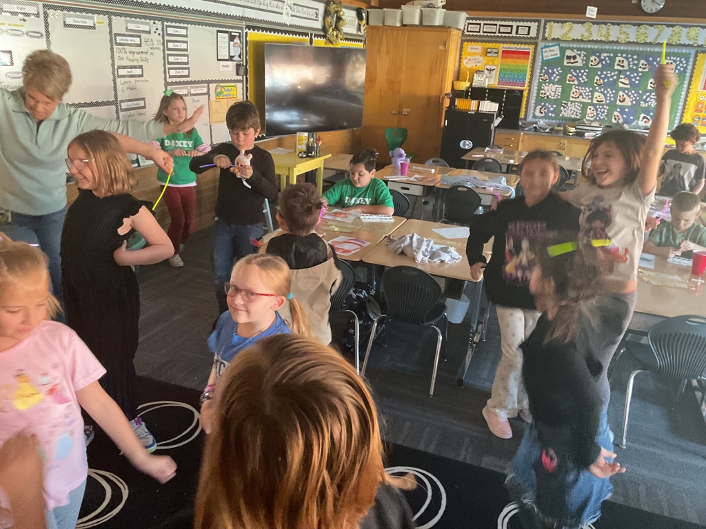 Students and a teacher dancing together in a classroom. One student in the foreground is cheering with a yellow glow stick raised high, while others are wearing "Doxey" green shirts.